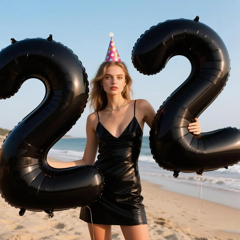 A young woman stands holding large black foil number balloons forming the number 22 against a beach backdrop.