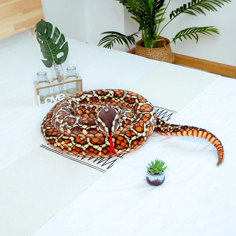A realistic, patterned snake plush toy is laid on a white surface next to a small succulent plant.