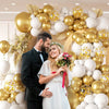A couple stands in front of a large balloon garland featuring white and gold balloons with confetti.