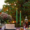 A vertical shot of green stick candles with lit flames set against a dark, leafy outdoor background.