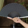 A hand is holding a black and white patterned folding hand fan with pleated ribs.