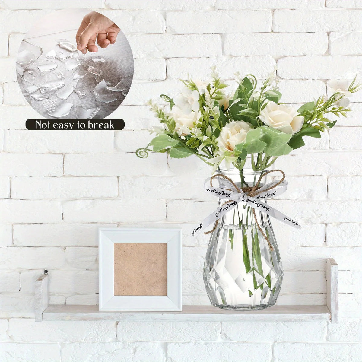 A clear faceted diamond crystal vase filled with white roses and greenery displayed on a white brick wall.