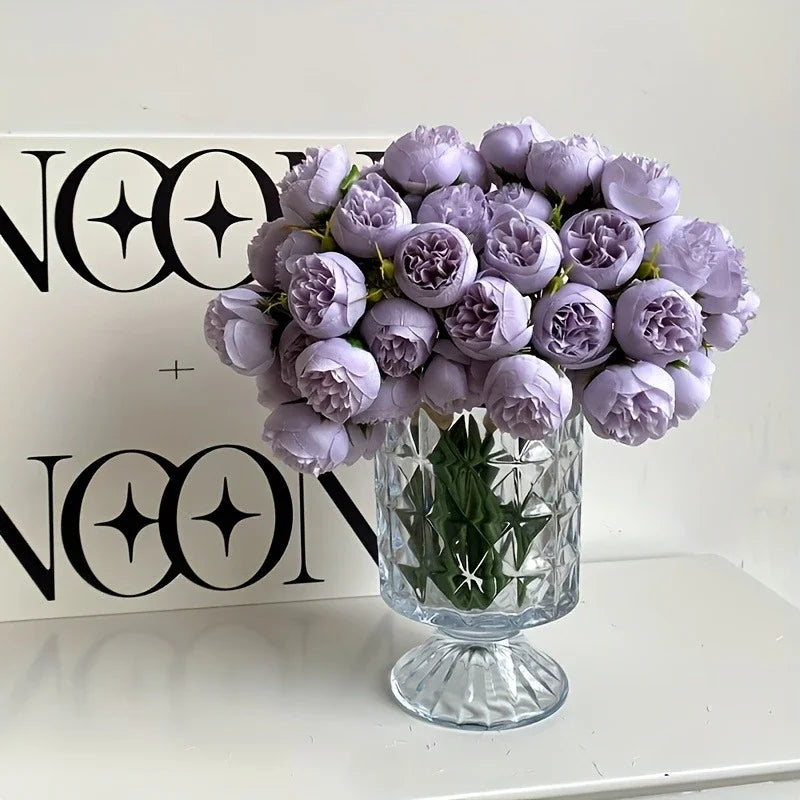 A bouquet of purple silk peonies in a glass vase displayed in front of a black and white logo box.