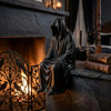 A black hooded figure resembling a grim reaper sits on a stone mantelpiece next to a fireplace and old books.