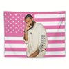 Un homme à la barbe portant une chemise de couleur claire pose devant un fond de drapeau américain rayé rose et blanc.