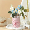 A handmade floral bouquet featuring light blue and white flowers in a pink ceramic vase on a white speckled plate.