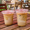 Two iced coffee drinks in clear plastic cups with lids and straws sitting on a wooden table.