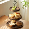 A wooden 3-tier cupcake stand displaying various baked goods like bread rolls and cupcakes.