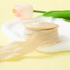 A roll of light beige decorative fishtail yarn ribbon resting on a white surface with a pink tulip in the background.
