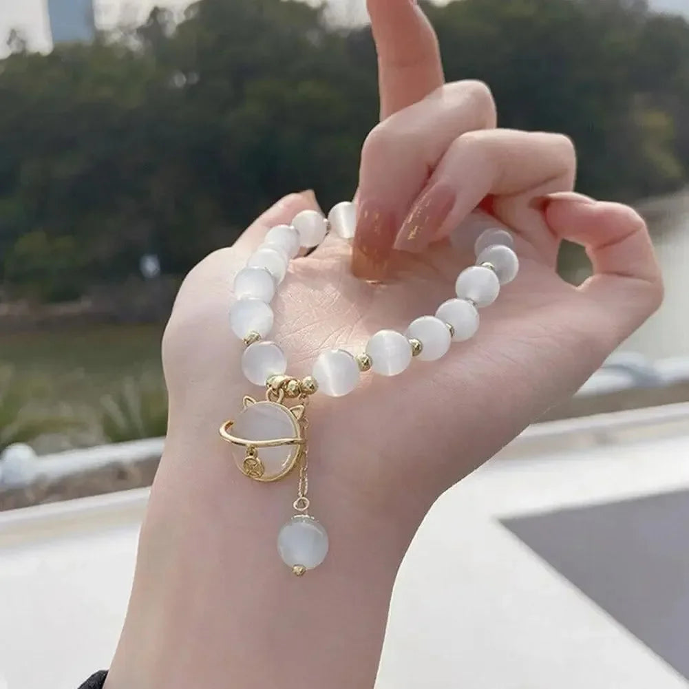 A hand is holding a white natural stone lily of the valley bracelet with a gold charm against a blurred outdoor background.