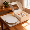 A white cardboard box with a lid is open, displaying a round cake board and several stacked round cake boards on a wooden tab