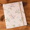 A beige linen photo album with delicate green and pink floral embroidery rests on a wooden surface.