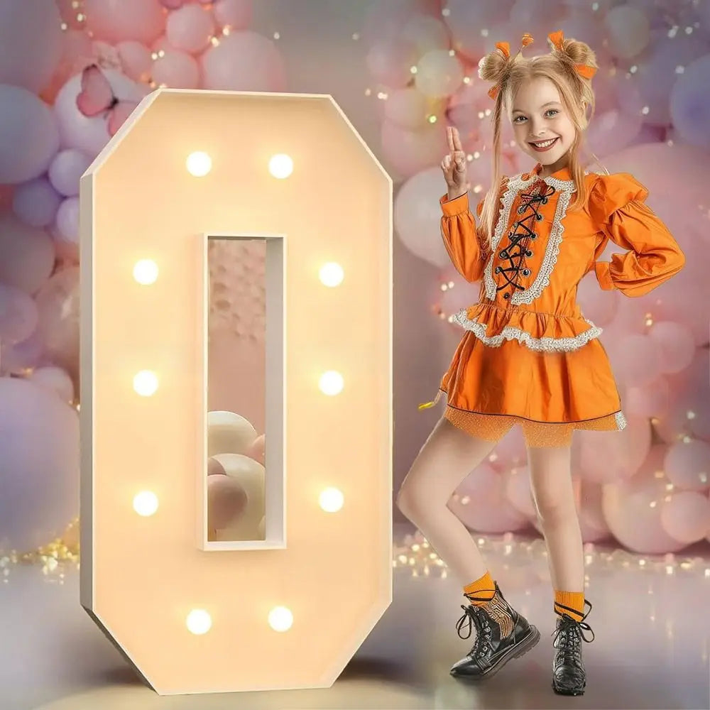 A young girl in an orange dress stands next to a glowing light-up number balloon decoration.