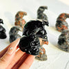 Une main tient un crâne de corbeau en obsidienne polie, décor entouré de sculptures en pierre sombre.