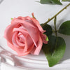 A pink velvet artificial rose with a green stem and leaves is displayed on a white surface.