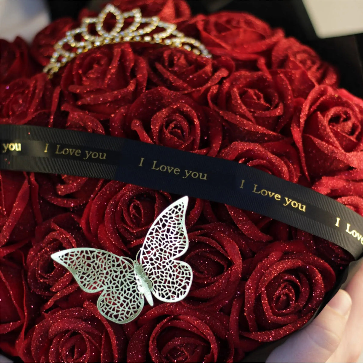 A bouquet of deep red glitter artificial roses with a silver butterfly decoration and a black ribbon that reads I Love you