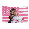 A photo of a male K-pop idol making a peace sign in front of a pink and white American flag tapestry.