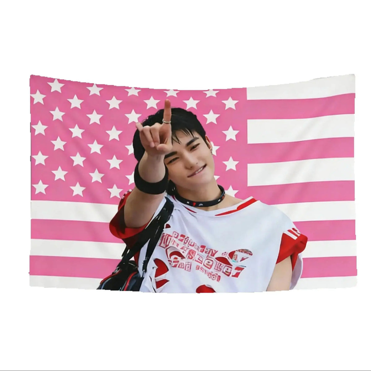 A photo of a male K-pop idol making a peace sign in front of a pink and white American flag tapestry.