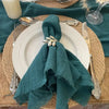 A stack of ten handcrafted fringed cotton napkins in a deep teal color displayed on a wooden surface.