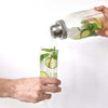 A hand pours liquid from a glass jar into a tall glass, showing a stainless steel shaker lid being used for seasoning or cock