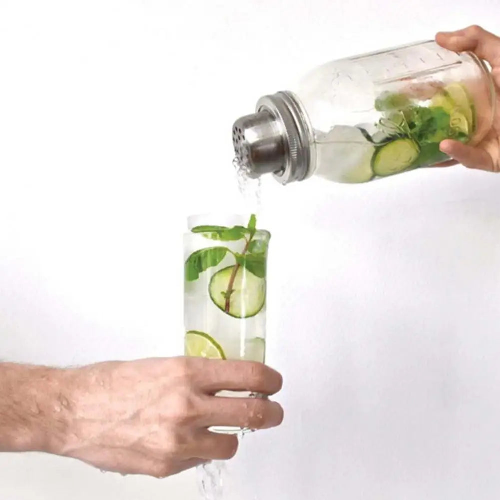 A hand pours liquid from a glass jar into a tall glass, showing a stainless steel shaker lid being used for seasoning or cock