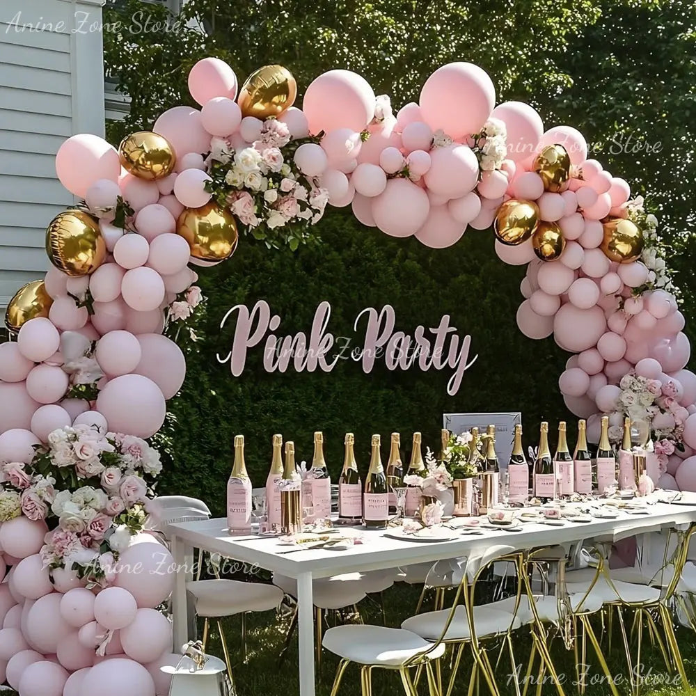 A floral arch made of pink latex balloons decorated with greenery and gold accents for a party backdrop.