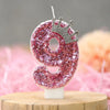 A pink sequin number nine candle with a silver crown topper displayed on a wooden surface.