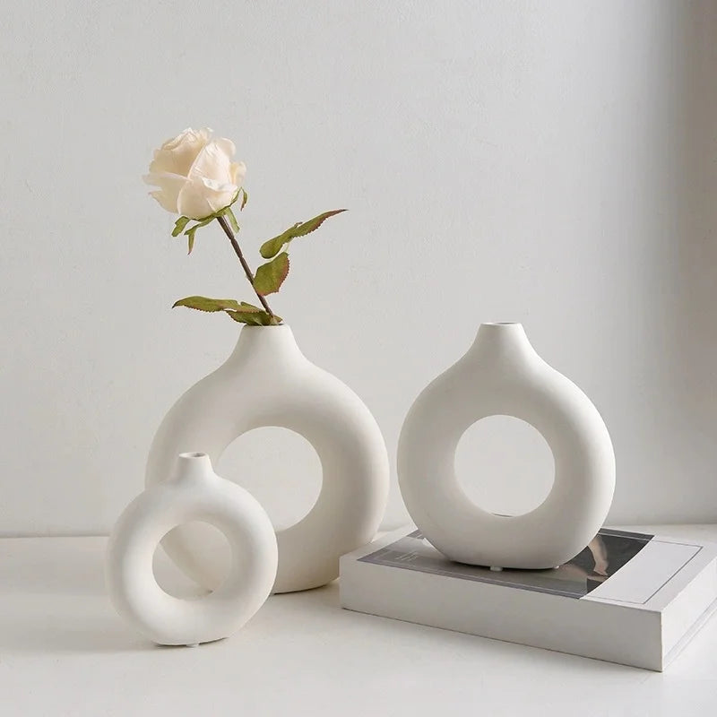 A white ceramic vase shaped like a donut holds a single pale pink rose against a light background.