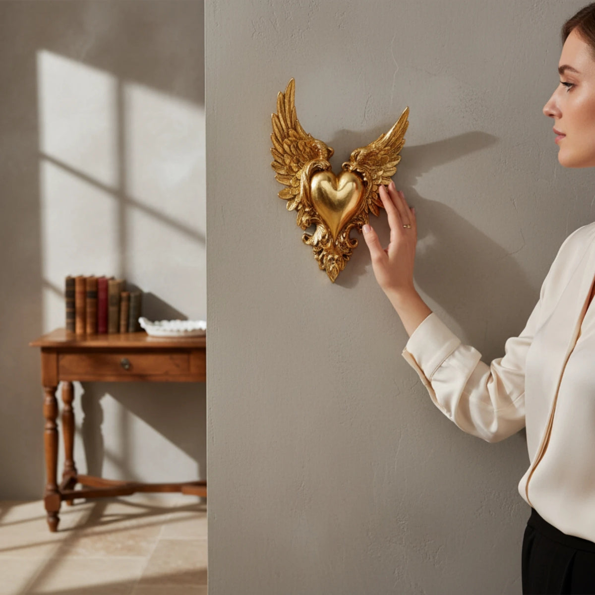 A woman's hand is placing an ornate gold winged heart wall decor on a gray wall.