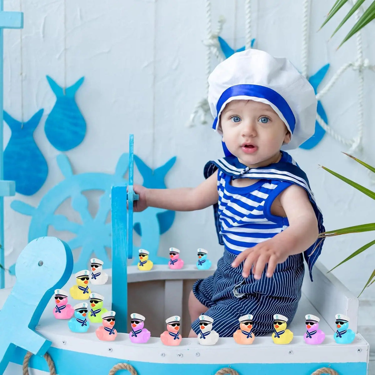 A baby wearing a chef hat is holding mini resin cruise ducks with sunglasses on a wooden surface.