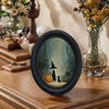 Oval wall art featuring a witch silhouette and a black cat in a misty woodland scene.