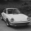 Une Porsche 911 blanche garée sur une route goudronnée avec de la verdure en arrière-plan.
