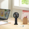 A black portable rechargeable handheld fan with a digital display is shown on a wooden desk next to a laptop.