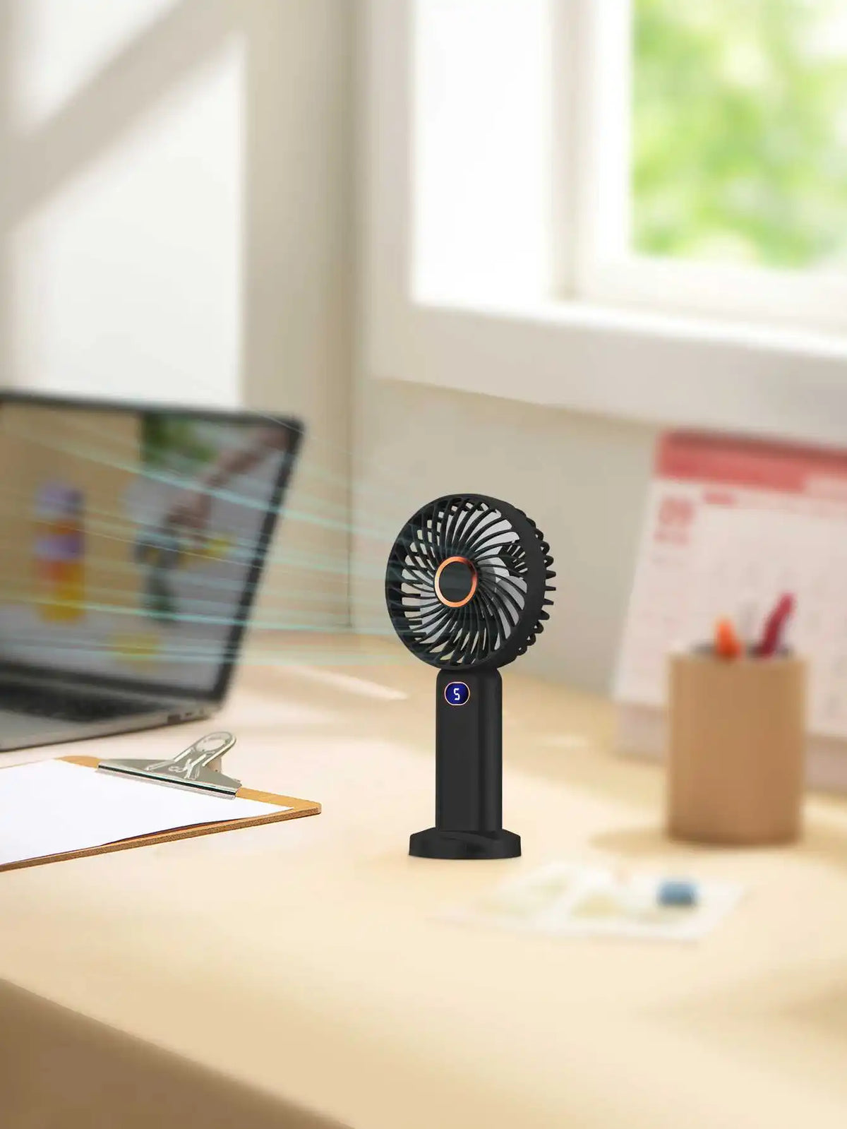 A black portable rechargeable handheld fan with a digital display is shown on a wooden desk next to a laptop.