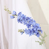Un délicat jet de fleurs de delphinium bleues artificielles avec des tiges vertes sur un fond de rideau blanc.