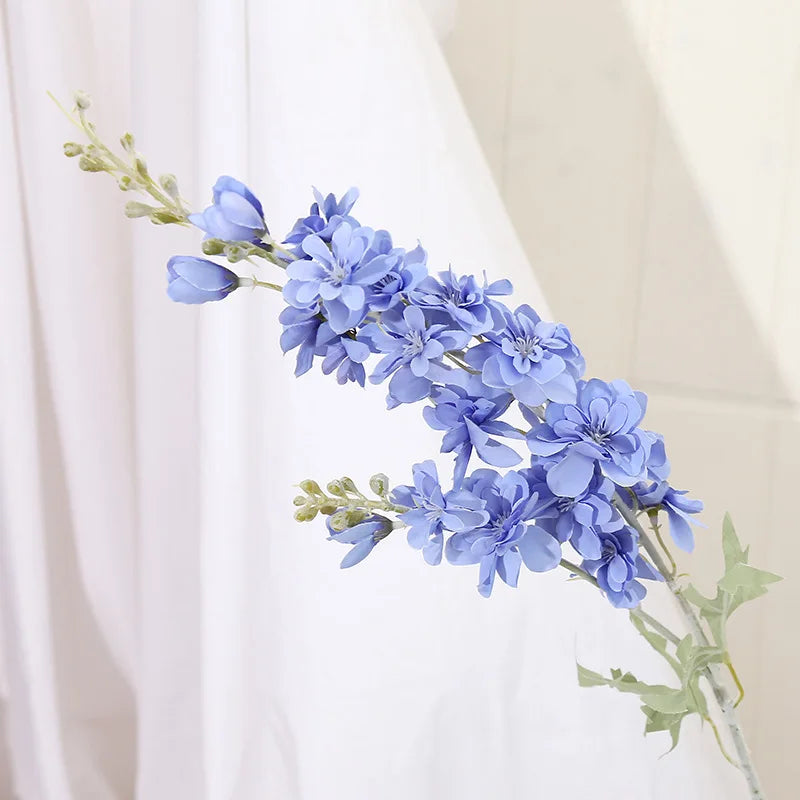 Un délicat jet de fleurs de delphinium bleues artificielles avec des tiges vertes sur un fond de rideau blanc.