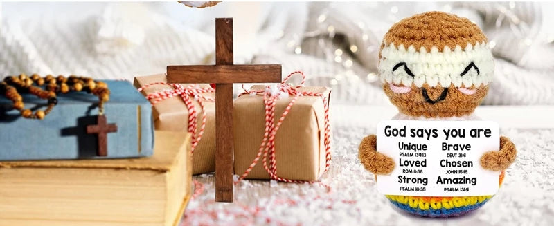 A brown crochet doll with a white headband and an encouragement card sits in front of a wooden cross and wrapped gifts.