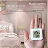 A hand holds a white digital thermometer and hygrometer device in a bedroom setting.