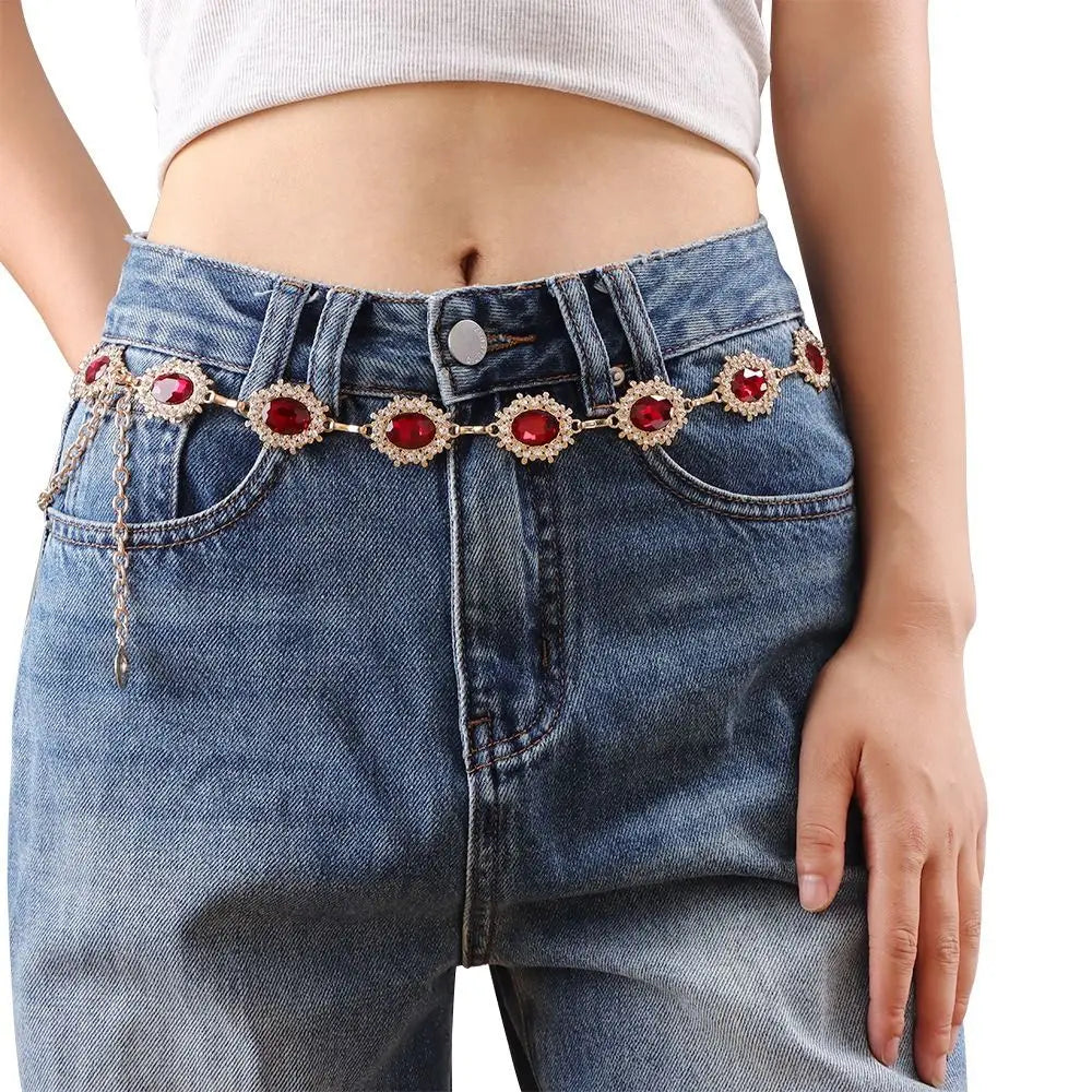 A close-up of denim jeans featuring a red crystal waist chain detail.