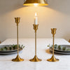A set of three matte gold taper candle holders with lit candles on a white tablecloth.