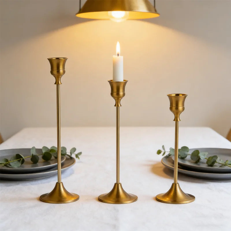A set of three matte gold taper candle holders with lit candles on a white tablecloth.