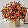 A vibrant bouquet of artificial silk roses and dried flowers in shades of deep orange, rust, and burgundy, displayed in a tex