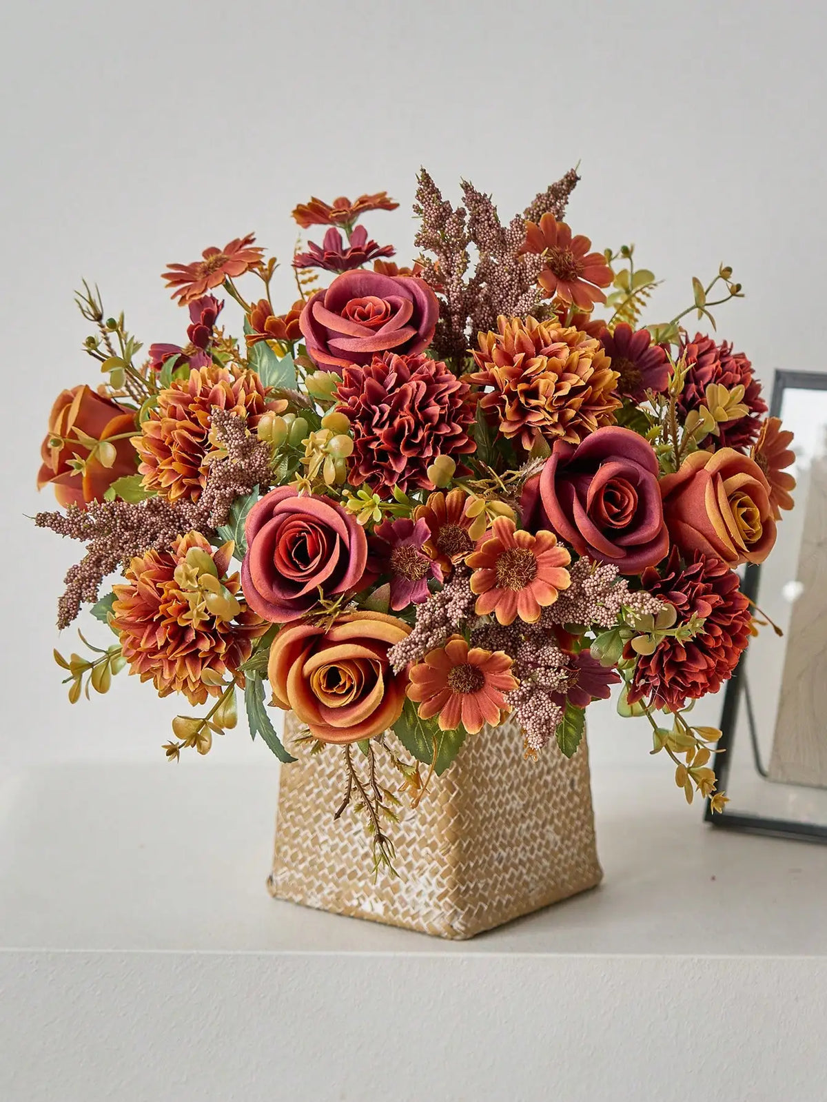A vibrant bouquet of artificial silk roses and dried flowers in shades of deep orange, rust, and burgundy, displayed in a tex
