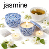 A collection of jasmine-themed soy wax candles, a lit candle in a blue patterned holder, and a glass cup of tea.