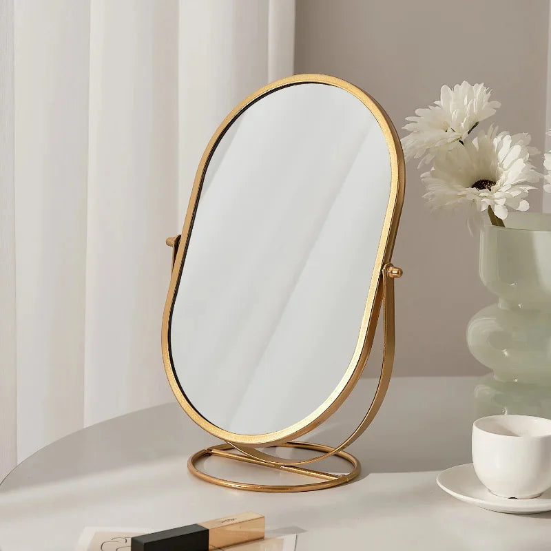 A gold-framed oval vanity mirror with a reflective surface sits on a white table next to a white cup and flowers.