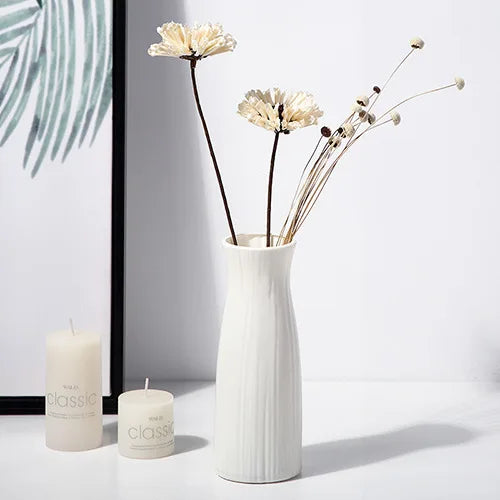A white ceramic vase holds dried white flowers and is displayed next to two pillar candles.