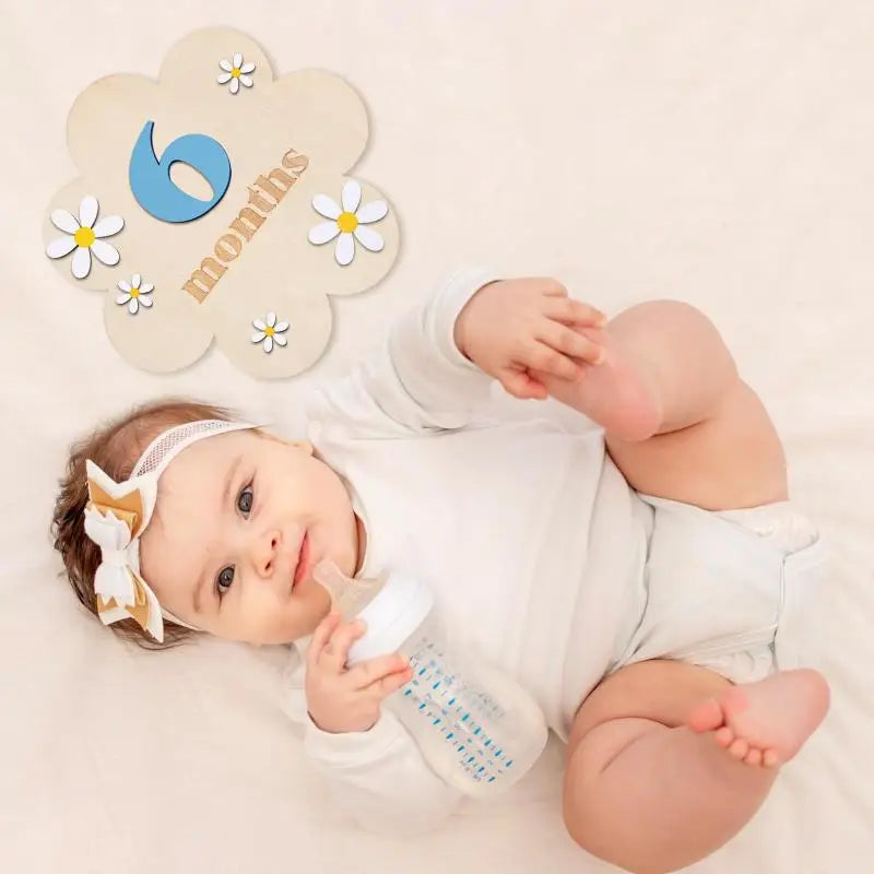 A baby lies on a white surface holding a milk bottle, with wooden monthly milestone cards nearby.