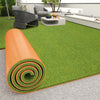 Rolled orange artificial grass carpet lying on a gray floor, with an outdoor living area in the background.