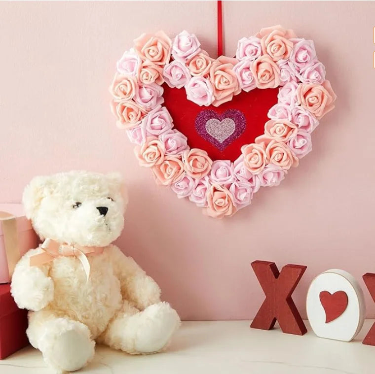A heart-shaped decoration made of pink and peach artificial foam roses hangs on a pink wall next to a white teddy bear.