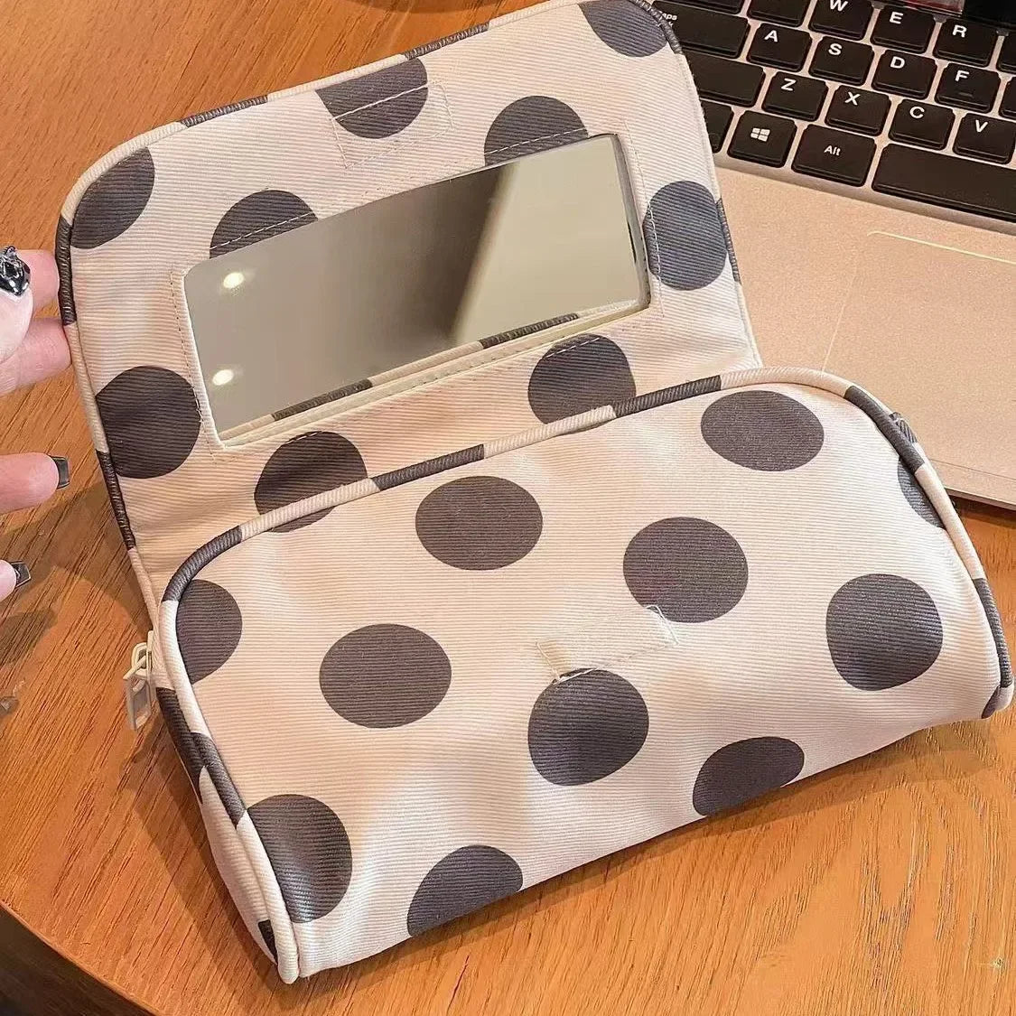 A beige canvas travel makeup bag with dark polka dots and a mirror, resting on a wooden surface next to a laptop.